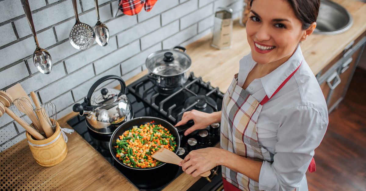 Woman smiling while cooking mixed vegetables on a stovetop in a cozy kitchen.