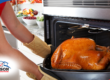 Person removing a golden roasted turkey from the oven, with a kitchen counter full of fresh produce in the background.